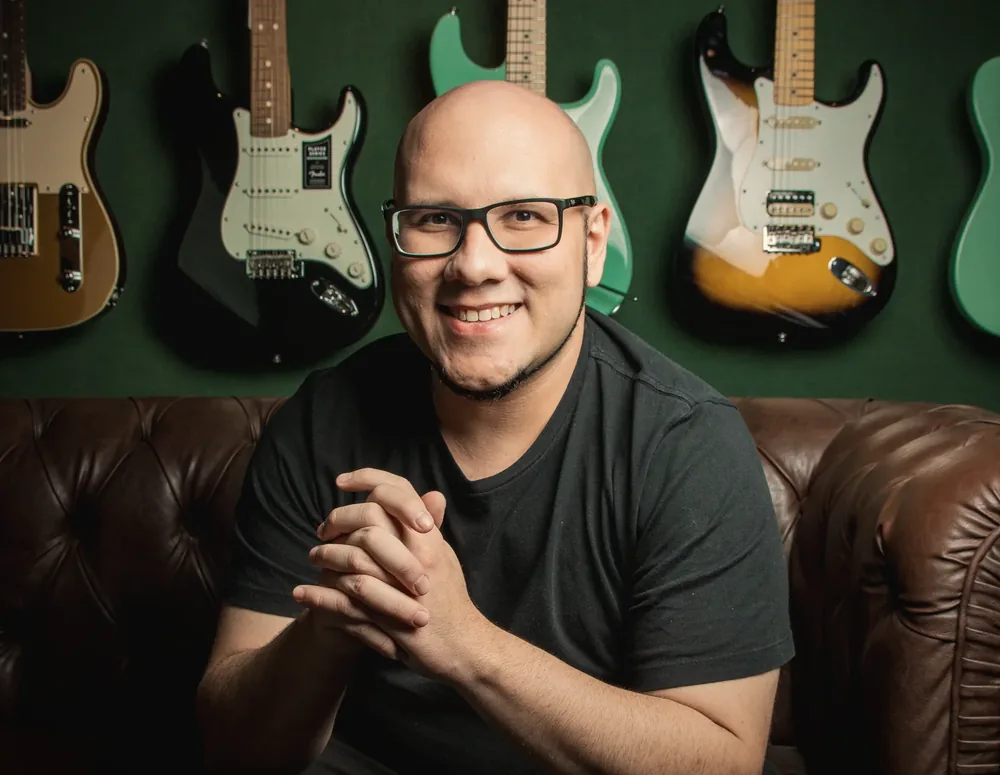 Daniel Parente sentado, sorrindo, com guitarras ao fundo